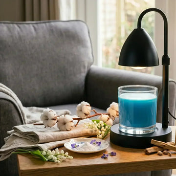 A blue wickless beeswax jar candle warmed under a black heat lamp on a wooden table, styled with raw cotton blossoms, linen cloth, white flowers, and sandalwood sticks next to a cozy armchair.