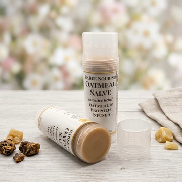 Two tubes of BaBee Nourish Oatmeal Salve are displayed on a light wooden surface with dried propolis and oatmeal and a cloth, against a soft, blurred background of white flowers.