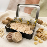 A black metal tin of honeycomb-shaped lotion rubs with a clear lid, shown open on a beige fabric surface. Two rubs are placed in front, and several cork and oat props surround the tin.