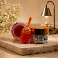 An elegant product photograph featuring a honey-themed lip mask set. A closed jar with a gold lid labeled 'LIP MASK DEEP REPAIR' (with a 'TB' logo) sits on a beige marble slab. Next to it is an open, tilted jar showing the red balm, and an orange honeycomb-patterned applicator wand leans between them. The background includes a vase with blurred baby's breath flowers and a lit brass table lamp near a dish of wax melts.