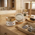 A detailed product shot on a wooden vanity features a jar of propolis-infused Gentle Grove Body Butter with a gold lid and botanical label. An open jar next to it contains the creamy grayish-beige butter. To the right, a tan leather valet tray holds a silver Bentley key fob, keys, and a stainless steel wristwatch. In the warm-lit, upscale bathroom background are a lamp, a bottle of Creed Green Irish Tweed cologne, and light wood cabinets with a mirror.