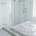A slatted rectangular shower steamer tray made of completely clear resin, resting on a wet white marble ledge. The modern, bright bathroom background features a glass-enclosed walk-in shower with white tile and chrome fixtures.