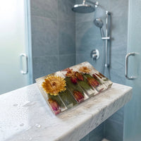 A slatted rectangular shower steamer tray made of clear resin containing embedded dried flowers, including a prominent yellow blossom and red rosebuds. The tray rests on a wet marble ledge with a modern tiled shower and silver fixtures in the background.