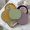 Three shower steamers in different colors (gold, green, purple, and lavender) with their foil wrappers, arranged on a white marble surface next to a folded white towel and a jar of bath salts.