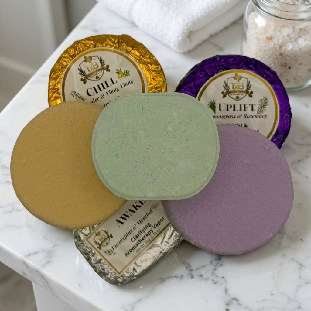 Three shower steamers in different colors (gold, green, purple, and lavender) with their foil wrappers, arranged on a white marble surface next to a folded white towel and a jar of bath salts.