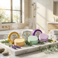 Three different varieties of shower steamers displayed on a white marble tray with water droplets and visible steam rising around them. From left to right: a gold foil-wrapped 