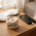 An open jar of Texas Bee Honeybee Body Butter resting on a light wood nightstand bathed in warm, natural sunlight. The jar displays a rich, creamy beige body butter with a swirled texture. Behind the product lies a white silk sleep mask, and to the right is a black smartphone plugged into a charging cable that hangs off the table's edge. A softly blurred bed with textured neutral linens is visible in the background.