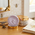 A detailed product photo on a light wood vanity counter shows an open and a closed jar of lavender body butter with polished gold lids. The open jar reveals a thick, light purple cream with a swirled texture, and its lid rests beside it. The label on the closed jar reads 'TXB LAVENDER BODY BUTTER' and features small bee and botanical illustrations. The background includes a gold-framed mirror with a decorative gold bee attached, wallpaper with a subtle honeycomb pattern and small bee drawings, a stack of te