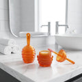 Two translucent orange, honeycomb-shaped jars of Texas Bee Lip Mask Deep Repair resting on a white marble vanity in a bright, modern bathroom. One jar is closed, while the other is open, displaying the lip mask inside with its pink-tipped applicator wand resting gently on the edge. In the softly blurred background, there are three rolled white hand towels, a modern faucet, and a white sink.