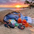 A woven beach bag rests on the sand at sunset with a blue and white striped towel spilling out. Scattered nearby are beach essentials including keys on a floral scrunchie, face wipes, spearmint gum, and two translucent orange, honeycomb-shaped jars of Texas Bee Lip Mask Deep Repair. One jar is closed, and the other is open with its applicator wand resting on the sand, illuminated by the warm, golden sunset over the ocean.
