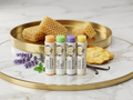 Four tubes of Texas Bee Propolis & Beeswax Lip Salve in Tropical Pineapple, Vanilla Mint, Lavender Lemonade, and Natural varieties lined up on a round gold tray, surrounded by natural ingredients including honeycomb, lavender sprigs, fresh mint leaves, a dried pineapple ring, and vanilla bean pods.