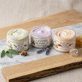 A product photograph featuring three open jars of Texas Bee body butter lined up on a white marble slab resting on a rustic wooden board. From left to right: Gentle Grove Body Butter with a pale green cream, styled with fresh sage leaves and a piece of propolis; Lavender Body Butter with a light purple cream, accompanied by sprigs of dried lavender; and Honeybee Body Butter with a soft peach cream, set next to a clear glass honey dipper and raw honeycomb. Each jar features a white label with gold trim and t
