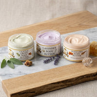 A product photograph featuring three open jars of Texas Bee body butter lined up on a white marble slab resting on a rustic wooden board. From left to right: Gentle Grove Body Butter with a pale green cream, styled with fresh sage leaves and a piece of propolis; Lavender Body Butter with a light purple cream, accompanied by sprigs of dried lavender; and Honeybee Body Butter with a soft peach cream, set next to a clear glass honey dipper and raw honeycomb. Each jar features a white label with gold trim and t