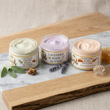 A product photograph featuring three open jars of Texas Bee body butter lined up on a white marble slab resting on a rustic wooden board. From left to right: Gentle Grove Body Butter with a pale green cream, styled with fresh sage leaves and a piece of propolis; Lavender Body Butter with a light purple cream, accompanied by sprigs of dried lavender; and Honeybee Body Butter with a soft peach cream, set next to a clear glass honey dipper and raw honeycomb. Each jar features a white label with gold trim and t