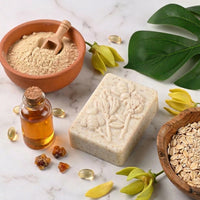 Rectangular handcrafted soap bar with an embossed floral and bee design, surrounded by a bowl of rolled oats, colloidal powder with a wooden scoop, a glass bottle of oil, and yellow blossoms on a marble surface.