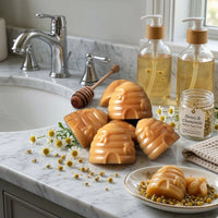 Handcrafted beehive-shaped honey and chamomile soaps arranged on a marble bathroom vanity with fresh chamomile flowers, a wooden honey dipper, and glass dispenser bottles.