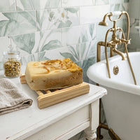 An open clear jar of smooth, creamy white Texas Bee body cream resting on a wooden nightstand. The cozy bedside setting includes a white lid, a plush sleep mask, an old book, an alarm clock, and a smartphone under warm lighting.