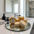 Round shower steamers on a circular mirror vanity tray on a white bathroom counter. Several steamers are wrapped in textured gold foil with a vintage Texas Bee label that reads 