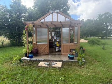 A wooden hobby greenhouse with a peaked roof and translucent panels sits on a concrete pad in a grassy yard. The double doors are swung open, revealing shelving with gardening supplies inside. A small wooden pergola-style overhang frames the front, decorated with potted plants on the concrete and a stone doormat featuring a bee design centered in front of the entrance.