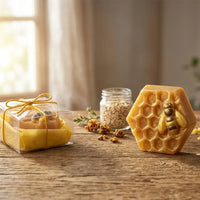 A golden, honeycomb-shaped Texas Bee Honey Oat Artisan Soap bar standing upright, featuring an embossed honeybee accented with shimmering gold details. To the left, a second bar is nestled inside a clear gift box lined with yellow paper and tied with a rustic twine bow. The artisanal soaps rest on a weathered wooden table, beautifully styled with scattered raw propolis chunks and a small glass jar filled with rolled oats in the softly blurred background near a sunlit window.