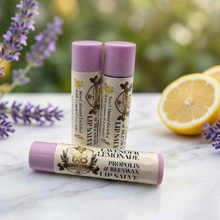Four tubes of Texas Bee Propolis & Beeswax Lip Salve in Tropical Pineapple, Vanilla Mint, Lavender Lemonade, and Natural varieties lined up on a round gold tray, surrounded by natural ingredients including honeycomb, lavender sprigs, fresh mint leaves, a dried pineapple ring, and vanilla bean pods.