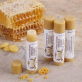 Four tubes of Texas Bee Natural Beeswax Propolis & Honey Lip Salve arranged on a textured stone surface. One tube in the foreground stands uncapped, showcasing the golden, natural balm inside. The remaining three tubes stand together in the background, displaying the brand's vintage-style label design. The product is beautifully styled with stacked pieces of raw, glistening honeycomb, small drops of yellow beeswax, and crumbled honeycomb bits scattered around the base.