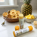 Two tubes of Texas Bee Pineapple Propolis & Beeswax Lip Salve displayed on a white countertop. One tube stands upright and uncapped, revealing a golden balm, while the other lies flat to show the product label. The tropical-themed scene is beautifully styled with a whole pineapple, a bowl of fresh pineapple chunks, a wooden bowl filled with dried pineapple rings, a bee-themed honey dipper, & a small glass dish of raw honeycomb against a white subway tile background.