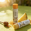 Two tubes of Pineapple Propolis & Beeswax Lip Salve from Texas Bee on a light green surface with warm, natural sunlight. One tube stands upright while the other lies horizontally in the foreground, showcasing a vintage-style label with a TXB logo. The tubes are accompanied by a fresh slice of pineapple, a small honey dipper with a puddle of golden honey, and scattered amber-colored propolis or honey granules.