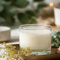 A Texas Bee PureBee Aromatic Wickless Vessel in the HoneyBee scent, featuring smooth cream-colored wax infused with pure honey fragrance oil inside a clear glass jar. The wickless vessel rests on a warm metallic tray alongside a delicate sprig of tiny yellow flowers, matches, and fresh eucalyptus leaves in a softly blurred, cozy setting.