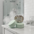 Two round, light green shower steamers with a textured finish rest on a white marble countertop. In the background, a third shower steamer stands upright, wrapped in crinkled silver foil with a vintage Texas Bee label that reads 
