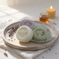 Three round Texas Bee Solid Lotion Bars embossed with an intricate honeybee design, arranged on a shallow wooden platter. The central, cream-colored HoneyBee bar sits prominently in front, flanked by a purple Quiet Breeze bar and a light moss green Gentle Grove bar, both resting in decorative paper liners. The platter is styled with dried lavender sprigs, while a small jar of honey and a lit beeswax candle glow softly in the warm, sunlit background.