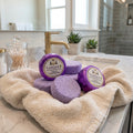 A stack of round, purple shower steamers resting on a soft, beige towel on a marble bathroom counter. Two of the steamers are wrapped in shiny purple foil with a vintage-style label from Texas Bee that reads 