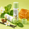 Two tubes of Vanilla Mint Propolis & Beeswax Lip Salve from Texas Bee on a light green background, surrounded by raw ingredients: fresh mint leaves, vanilla beans, white blossoms, a piece of honeycomb dripping with honey, and raw propolis chunks.