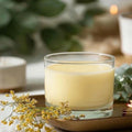 A Texas Bee PureBee Aromatic Wickless Vessel in the Garden Herb scent, featuring smooth cream-colored wax infused with pure honey fragrance oil inside a clear glass jar. The wickless vessel rests on a warm metallic tray alongside a delicate sprig of tiny yellow flowers, matches, and fresh eucalyptus leaves in a softly blurred, cozy setting.