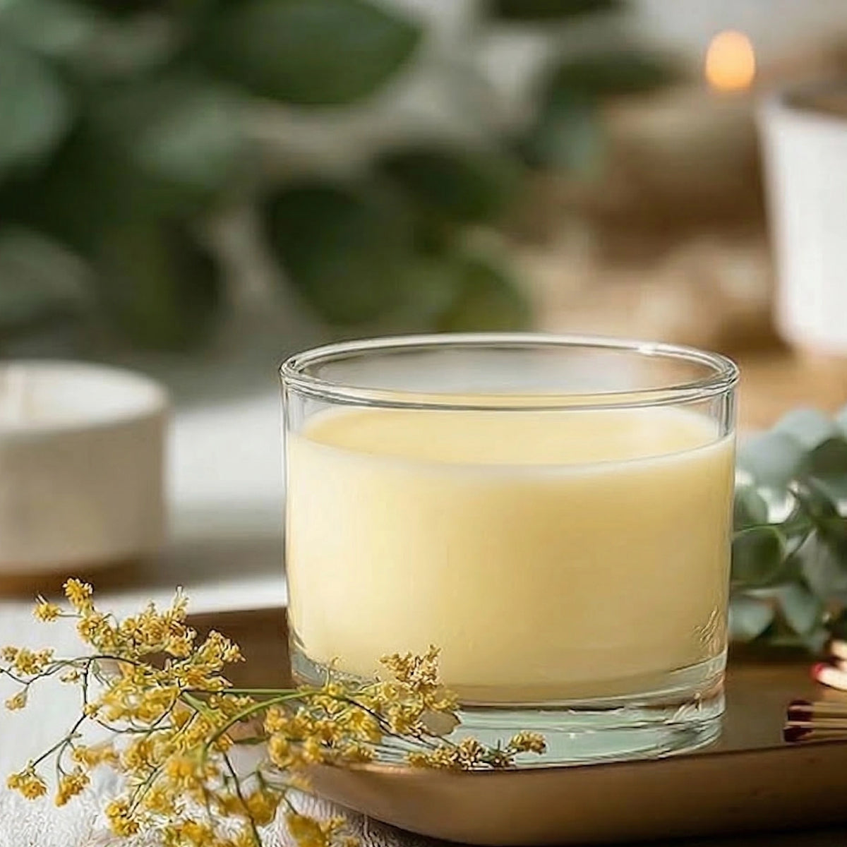 A Texas Bee PureBee Aromatic Wickless Vessel in the Garden Herb scent, featuring smooth cream-colored wax infused with pure honey fragrance oil inside a clear glass jar. The wickless vessel rests on a warm metallic tray alongside a delicate sprig of tiny yellow flowers, matches, and fresh eucalyptus leaves in a softly blurred, cozy setting.