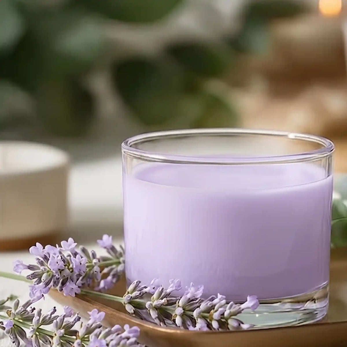 A clear glass jar candle filled with light lavender-colored wax, styled on a wooden tray with lavender sprigs against a softly blurred green and neutral background.
